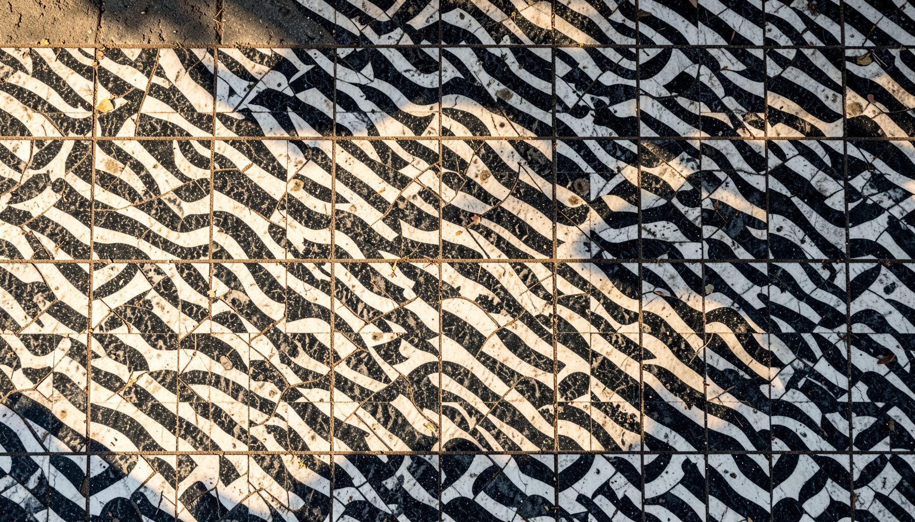 Overhead flat-lay photograph of a tiled Lisbon sidewalk with black-and-white wave patterns, warm sunlight casting long shadows.